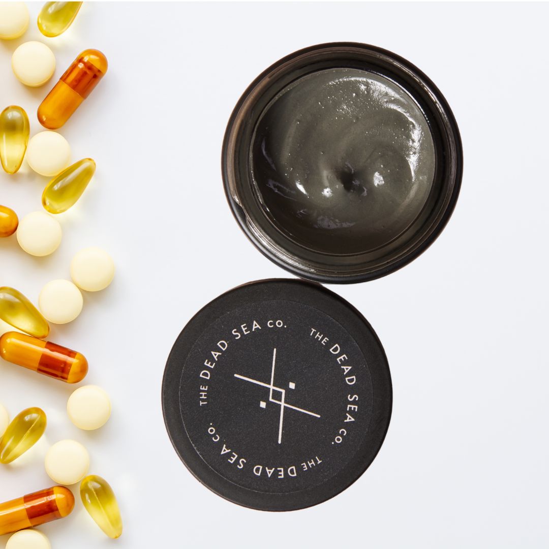 Flat lay image showing an open jar of Dead Sea mud mask next to a scattering of yellow and orange supplement capsules and tablets on a white background.