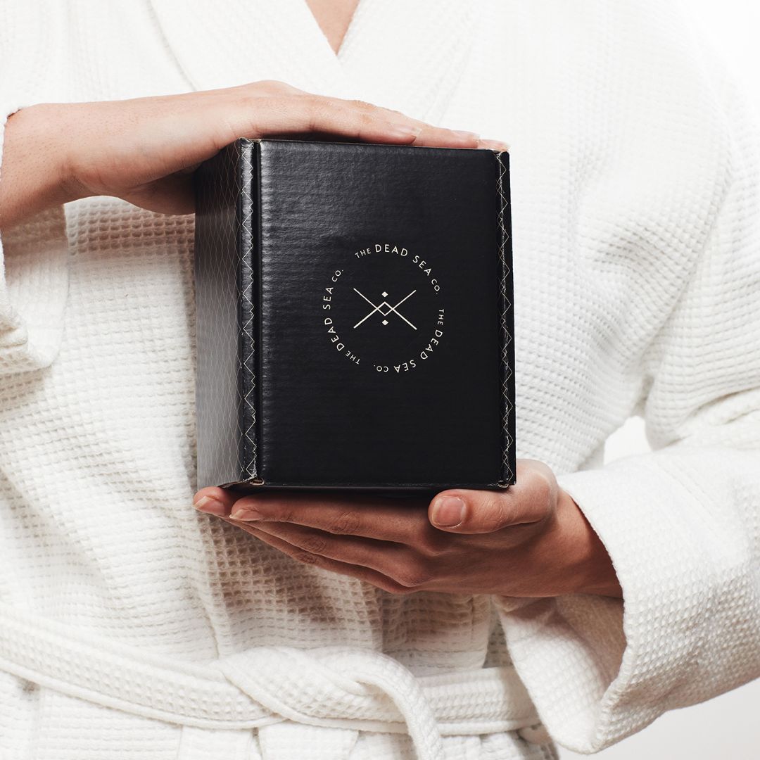 Person wearing a white waffle robe holding a black gift box from The Dead Sea Co., featuring their signature logo, evoking a sense of luxury and wellness.