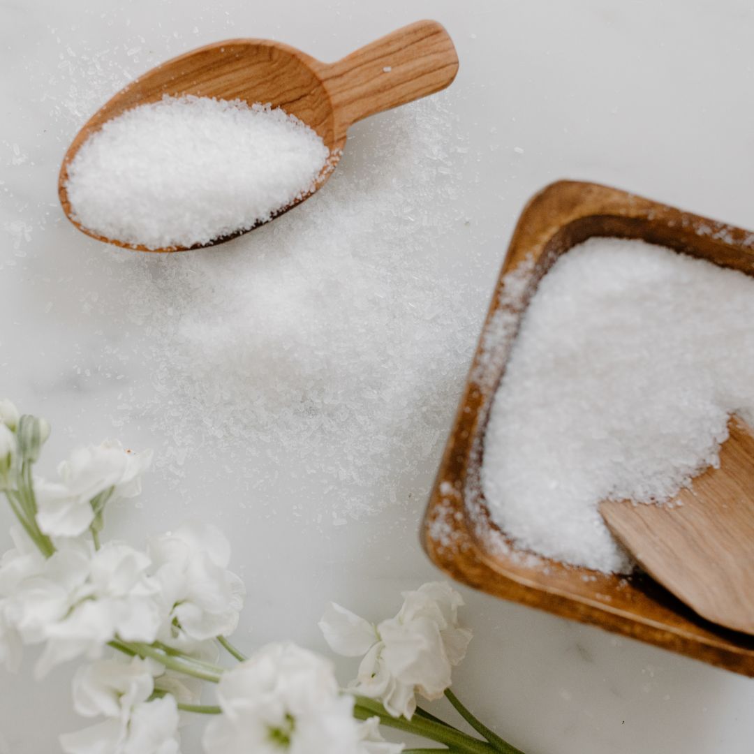A close-up of natural white bath salts in a wooden scoop and bowl, with white flowers on a marble surface, representing Dead Sea and Epsom salts used for skincare and relaxation.
