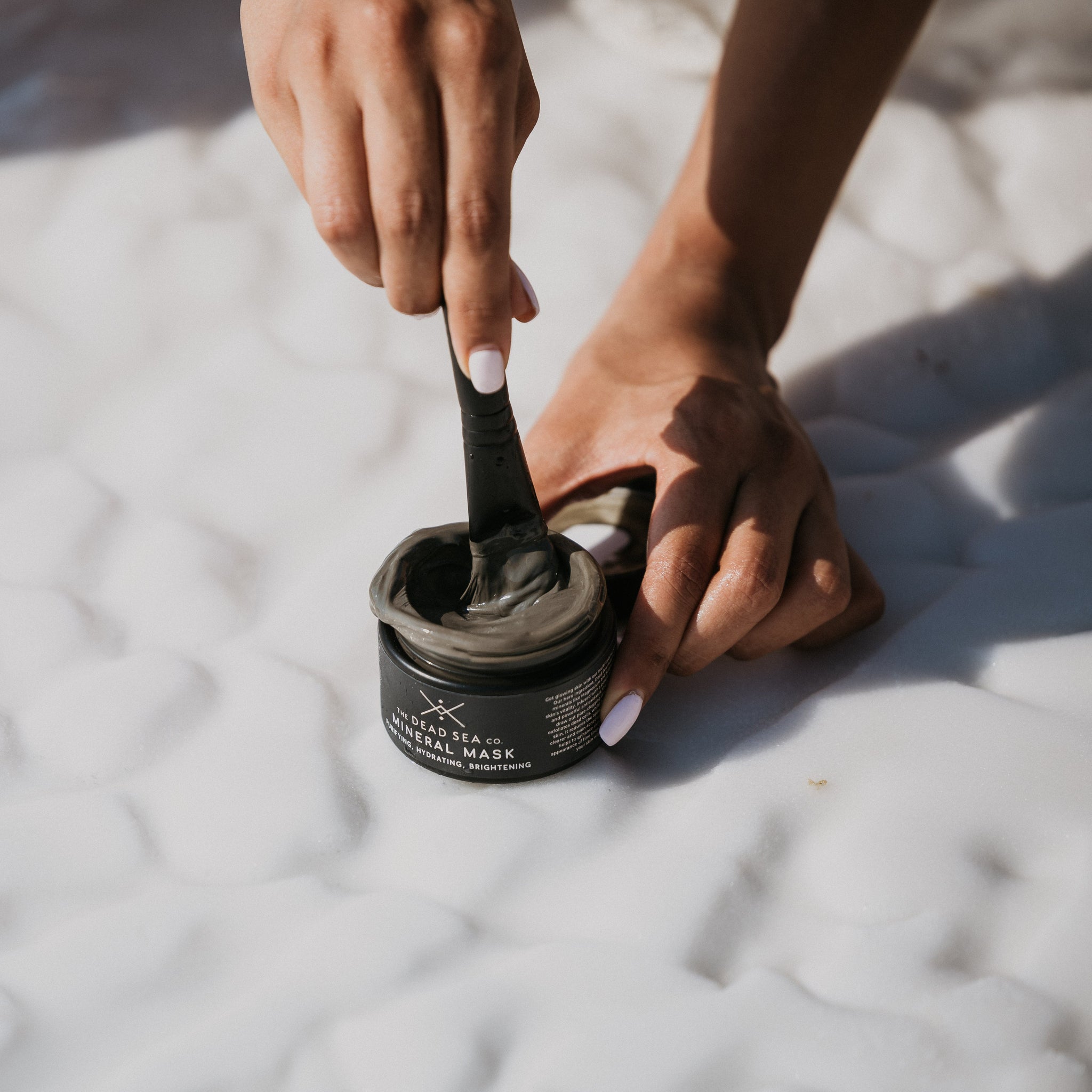 Hand stirring Dead Sea Mineral Mask with a brush, set against the shores of the Dead Sea.
