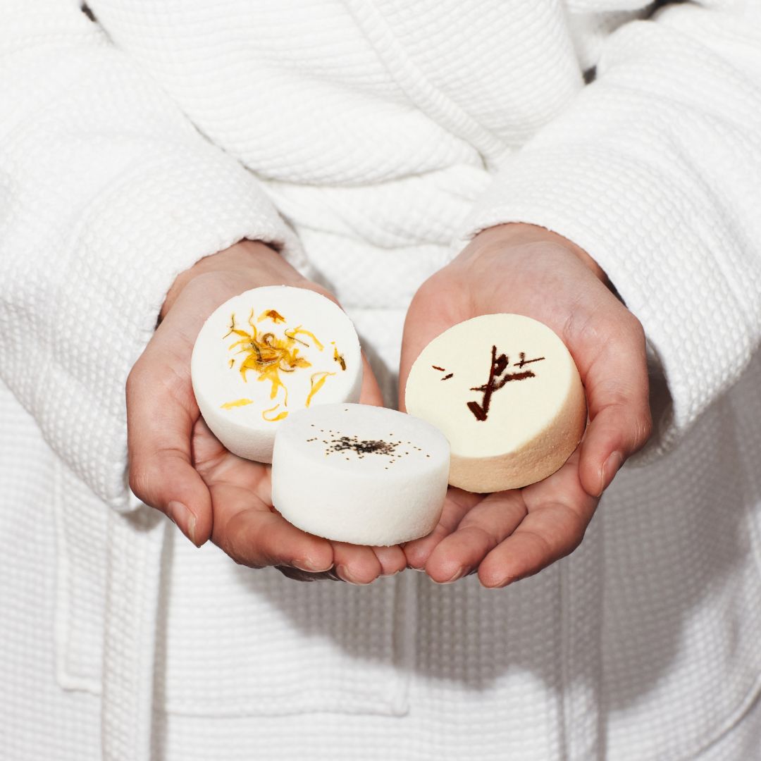Three handmade aromatherapy shower steamers with botanicals, elegantly held by a woman in a dressing gown, evoking a luxurious self-care ritual.