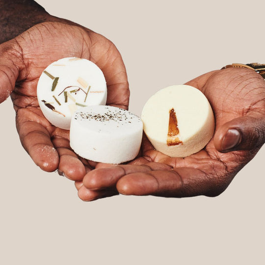 The image shows a man holding three aromatherapy shower steamers with unique botanicals placed on top. The first steamer is labeled 