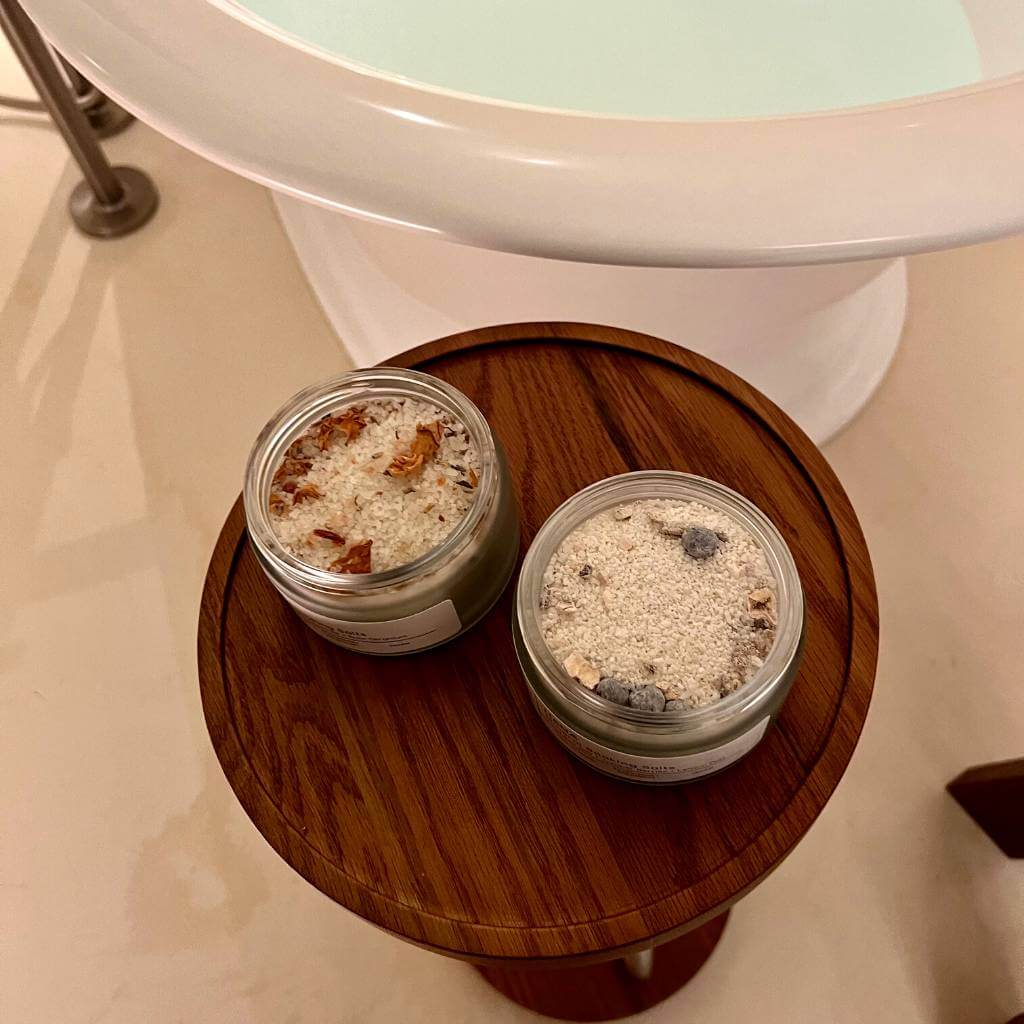 Dead Sea Bath Salt with dried botanicals and open lids sitting on table next to roll top bath