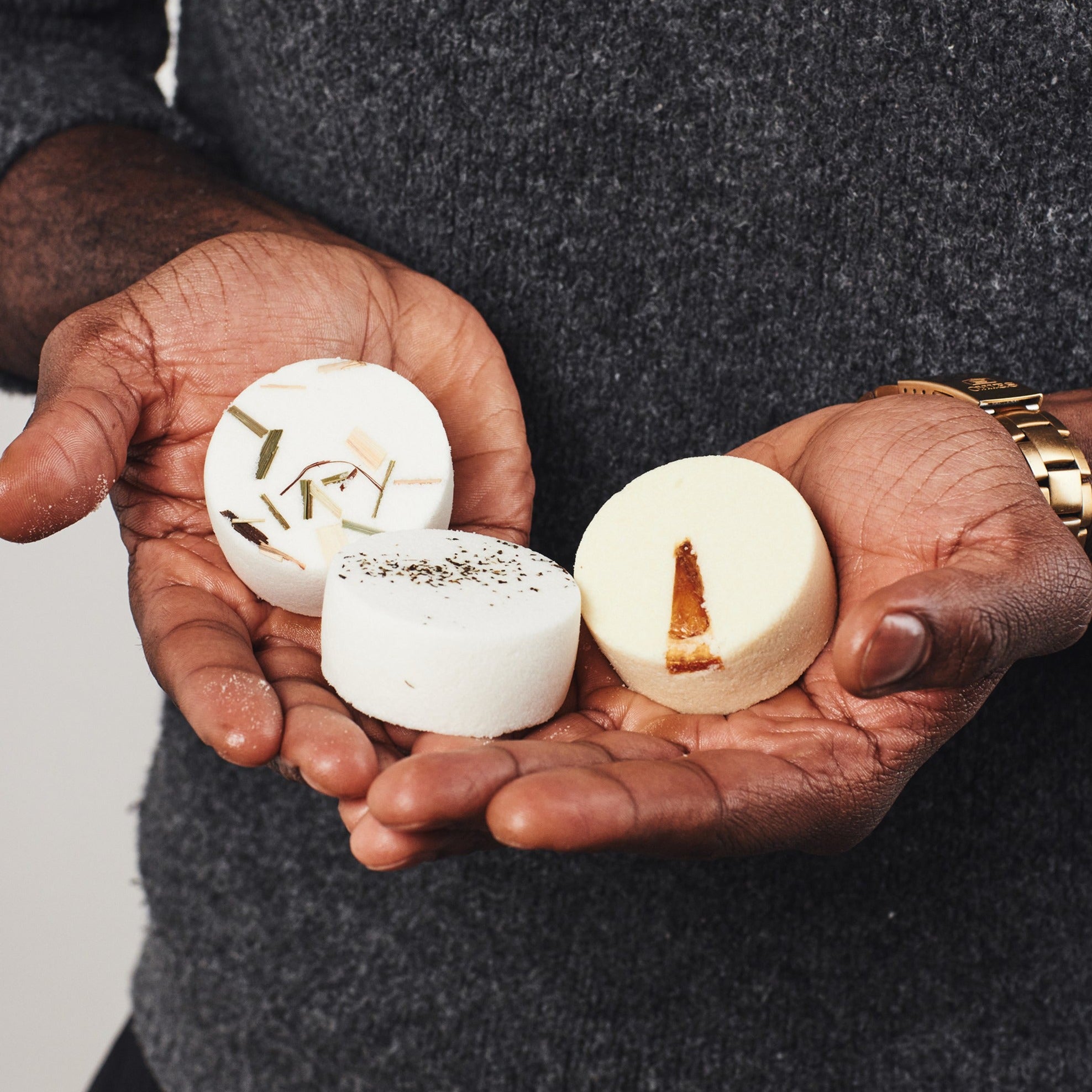 The image shows a man holding three aromatherapy shower steamers with unique botanicals placed on top. The first steamer is labeled 