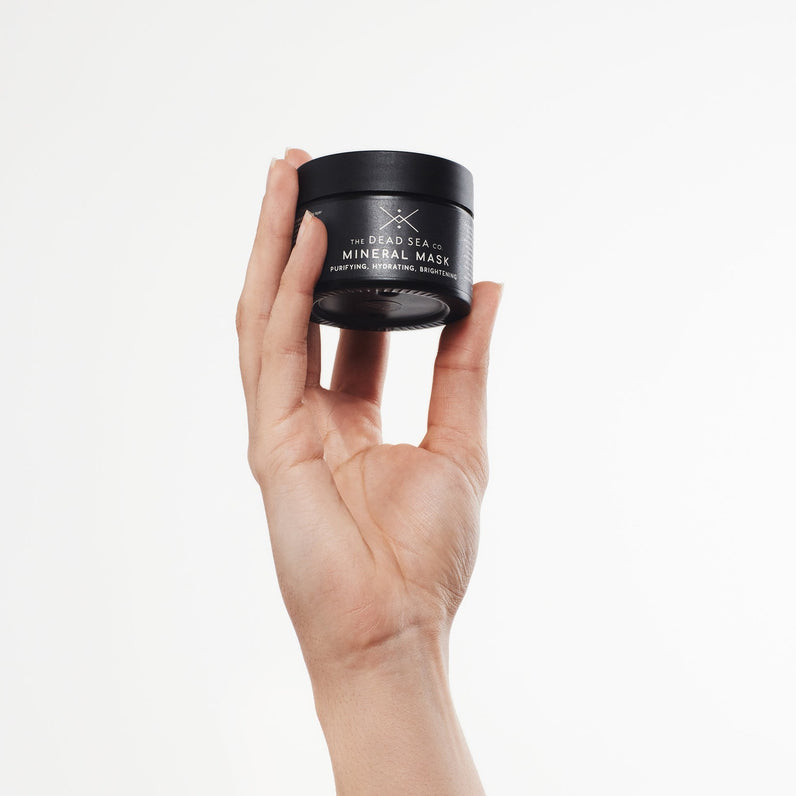 A woman's hand is holding a black glass jar containing the Dead Sea mud mask. The packaging has a sleek and elegant appearance, with a minimalist design that exudes a sense of sophistication. The jar is reflective, and the black color adds a touch of luxury to the overall aesthetic.