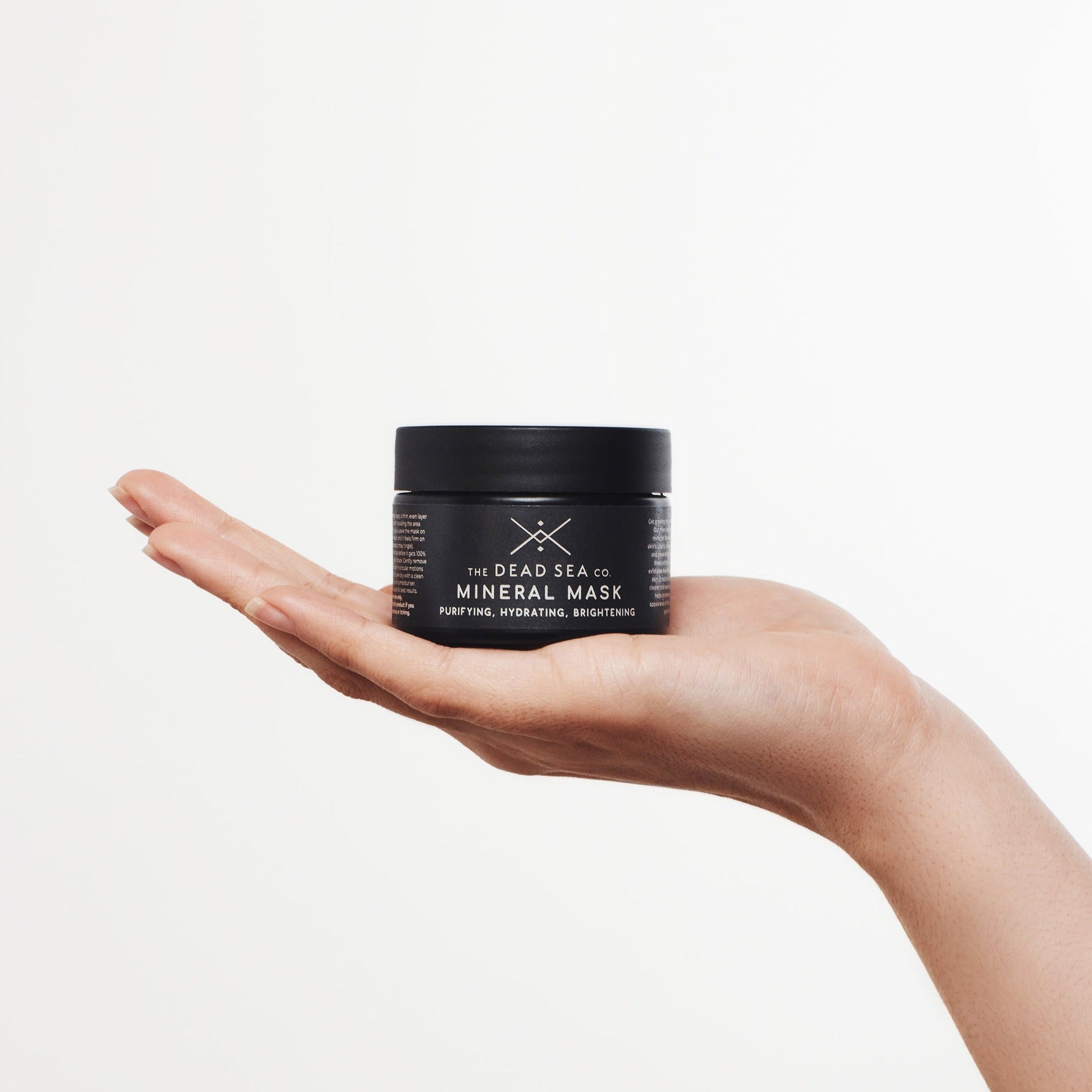 A woman's hand is gracefully holding a black glass jar, which houses the Dead Sea mud mask. The packaging has a sleek and sophisticated design, characterized by its minimalist style. The reflective jar exudes luxury, and its black color adds a touch of elegance to the overall aesthetic.